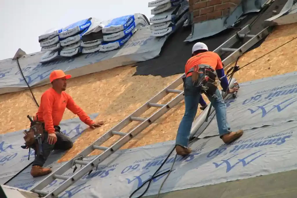 Two construction workers wearing safety gear installing roofing underlayment on a house roof.