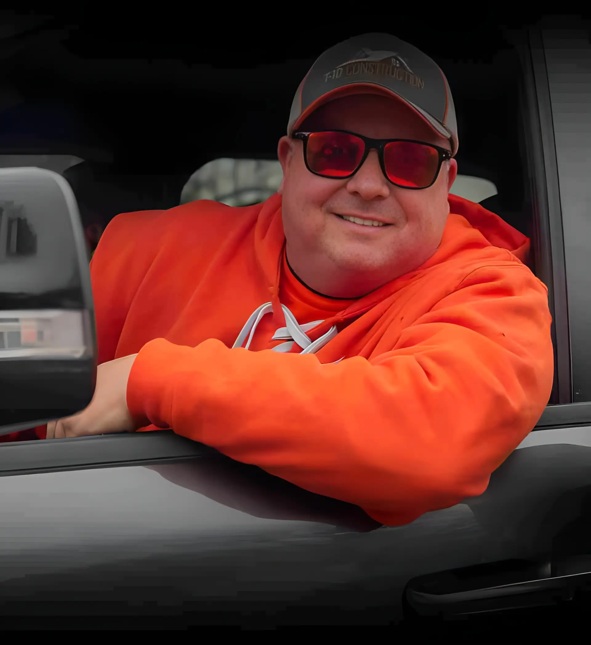 Man wearing a gray cap, red sunglasses, and orange hoodie leaning out of a black vehicle window.