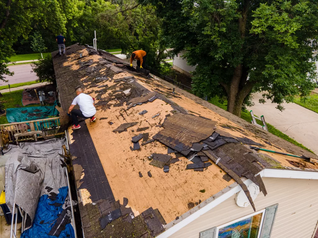 Three workers removing old shingles from a house roof with debris scattered on the surface.