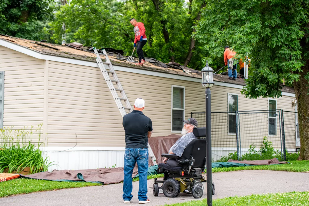 Workers repair the roof of a beige mobile home as two men watch from the ground outside.