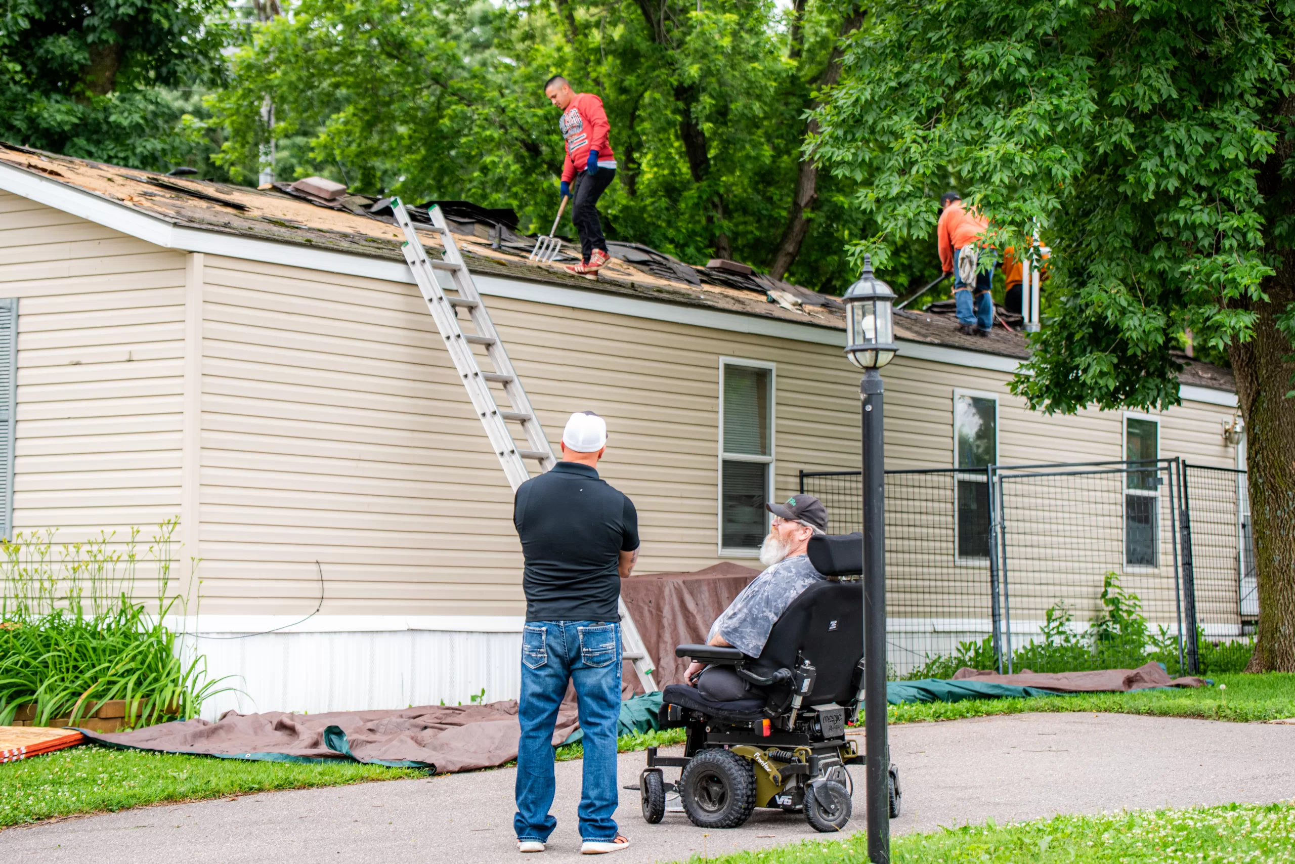 Workers repair the roof of a beige mobile home as two men watch from the ground outside.