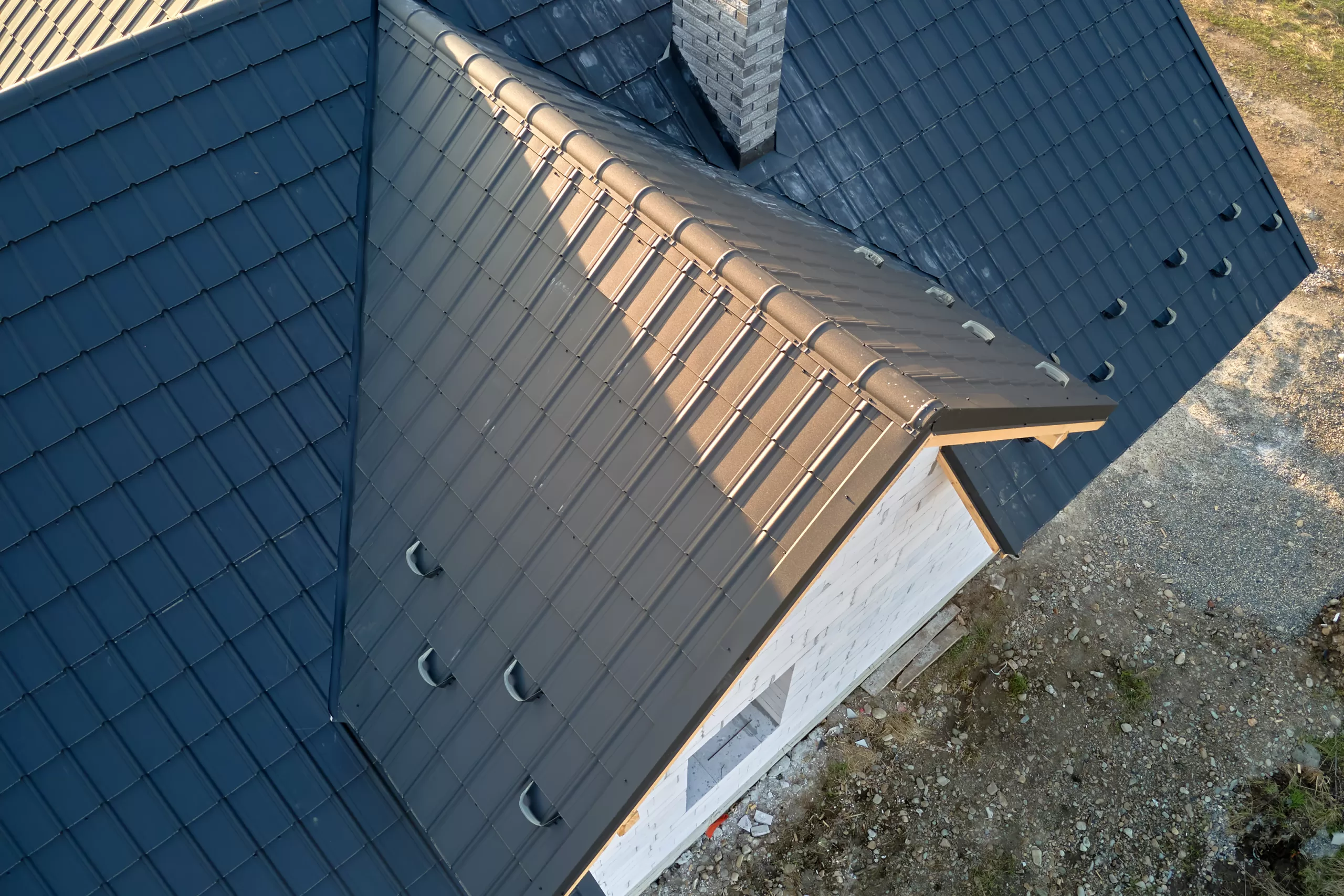 Aerial view of a house roof with black metal tiles, chimney, and surrounding gravel area.