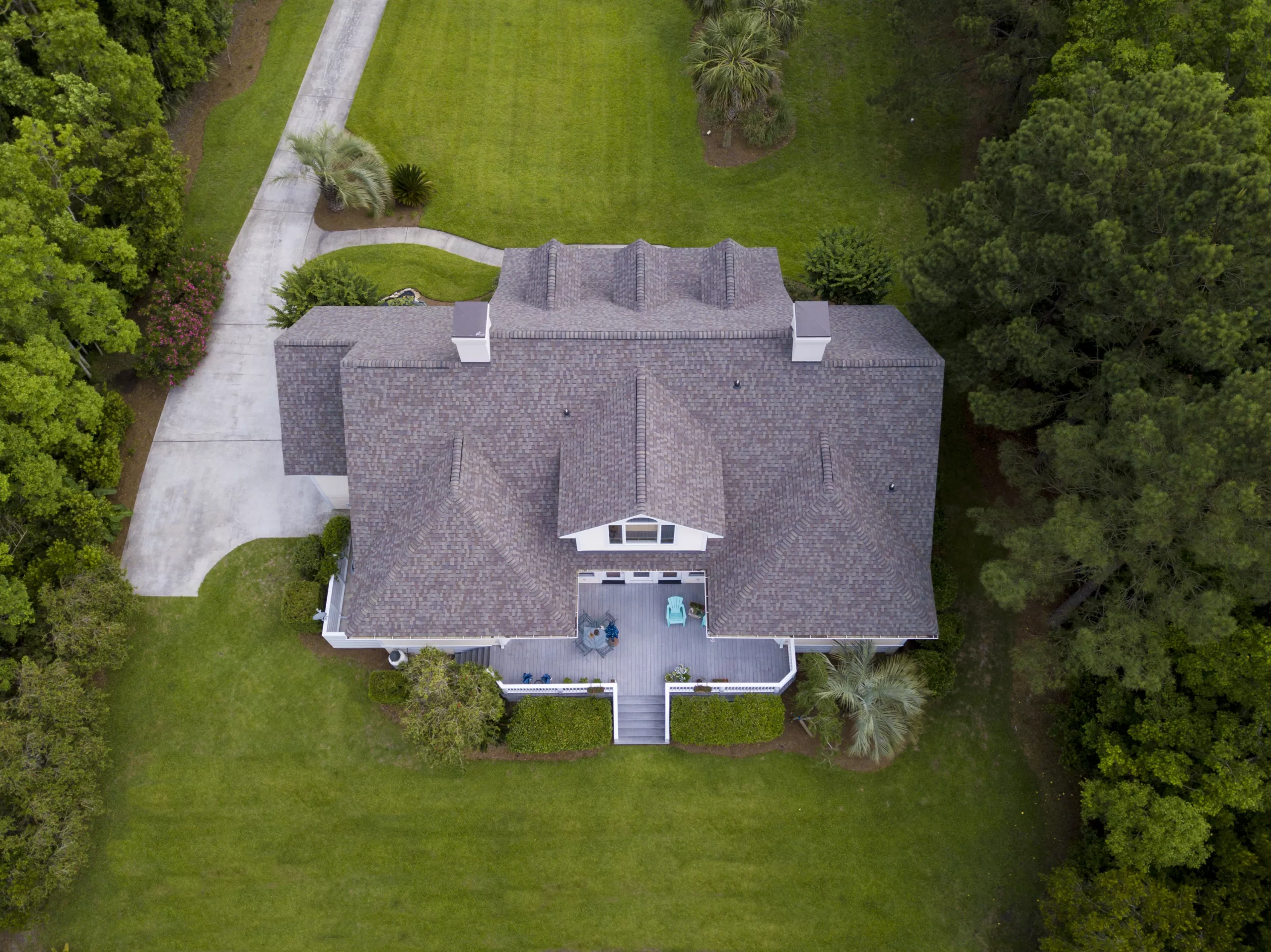 Aerial view of a house with gray roof, back porch, driveway, and surrounding green lawn and trees.