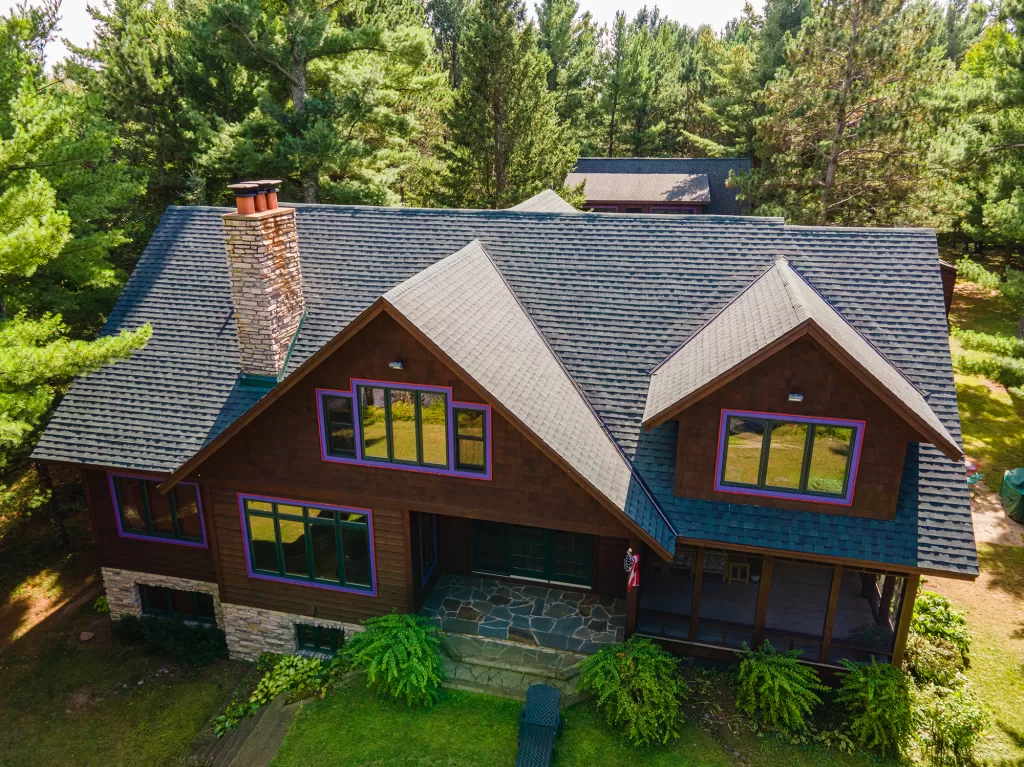 Two-story wooden house with dark shingled roof, stone chimney, large windows, and landscaped yard.