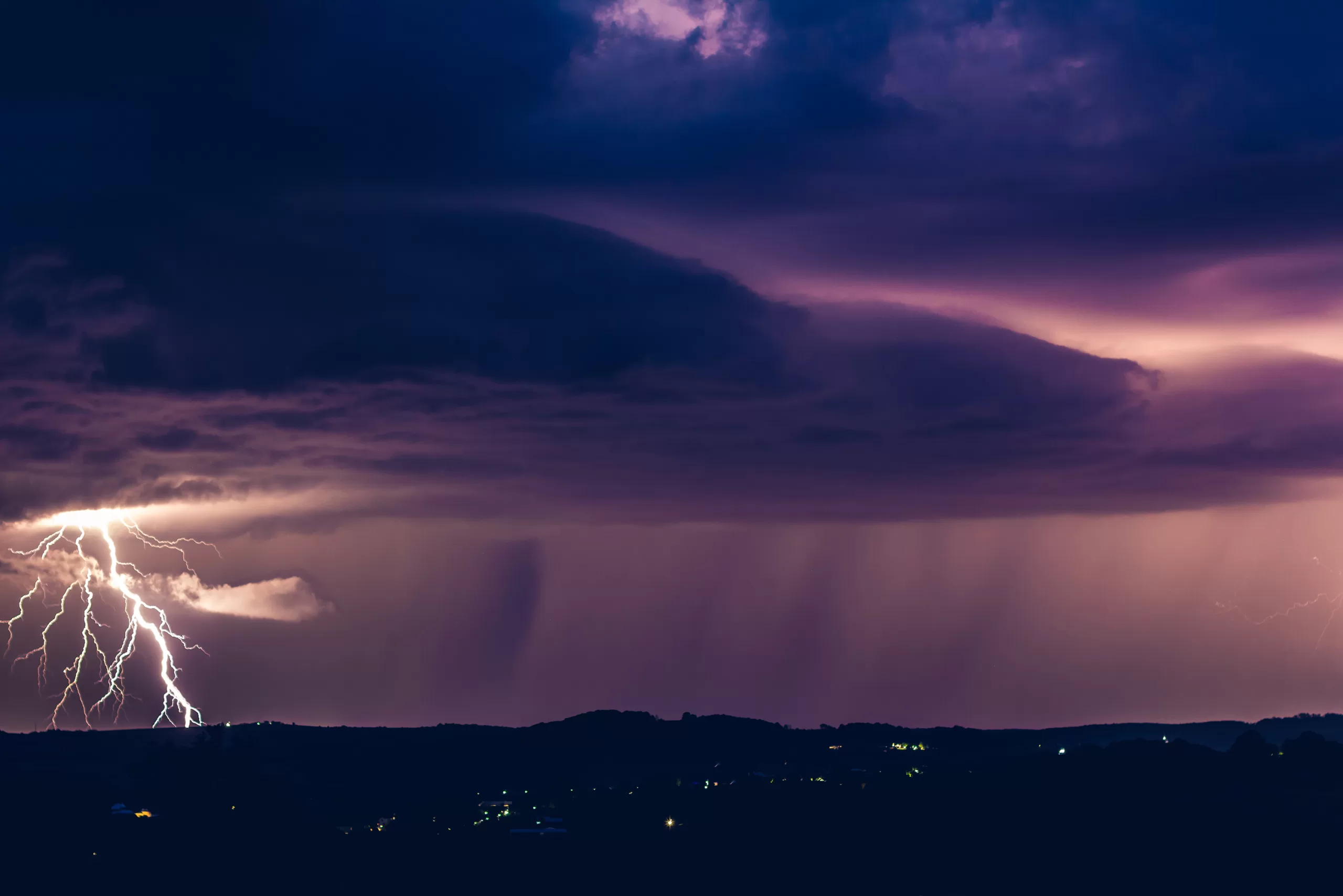 Nighttime storm with lightning striking left and rain falling over hills below.